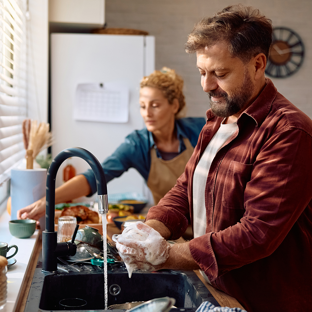 Smiling man washing dishes while cooking with his wife in the kitchen.