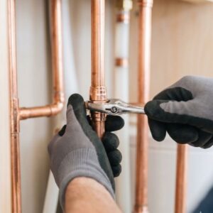 A professional plumber wearing grey work gloves using a wrench to adjust a copper water pipe.